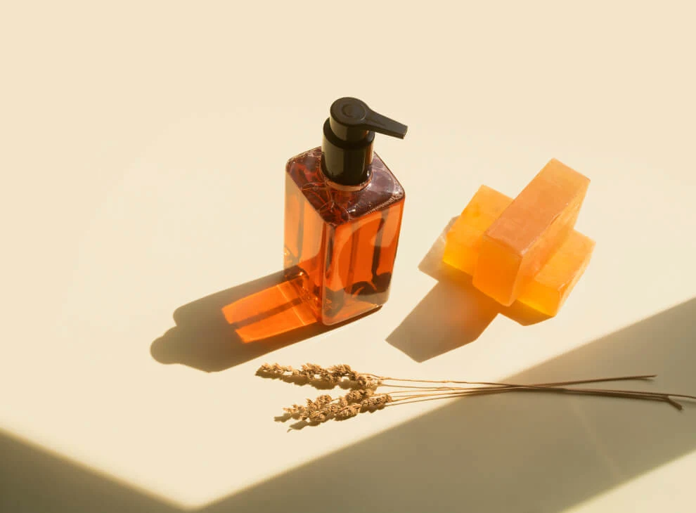 Un distributeur de savon liquide couleur ambre et deux barres de savon couleur abricot sur un fond pastel. Des fleurs séchées ont été disposées devant les savons.