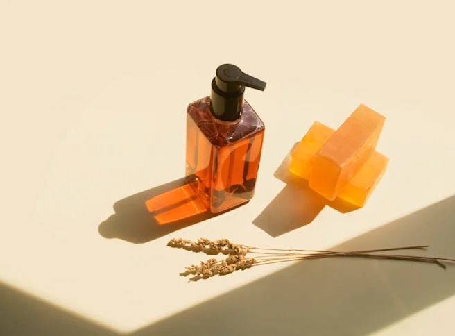 A bottle of amber-colored liquid soap and two bars of apricot-colored soap are on a pastel background. Dried flowers are displayed in front of the soaps.