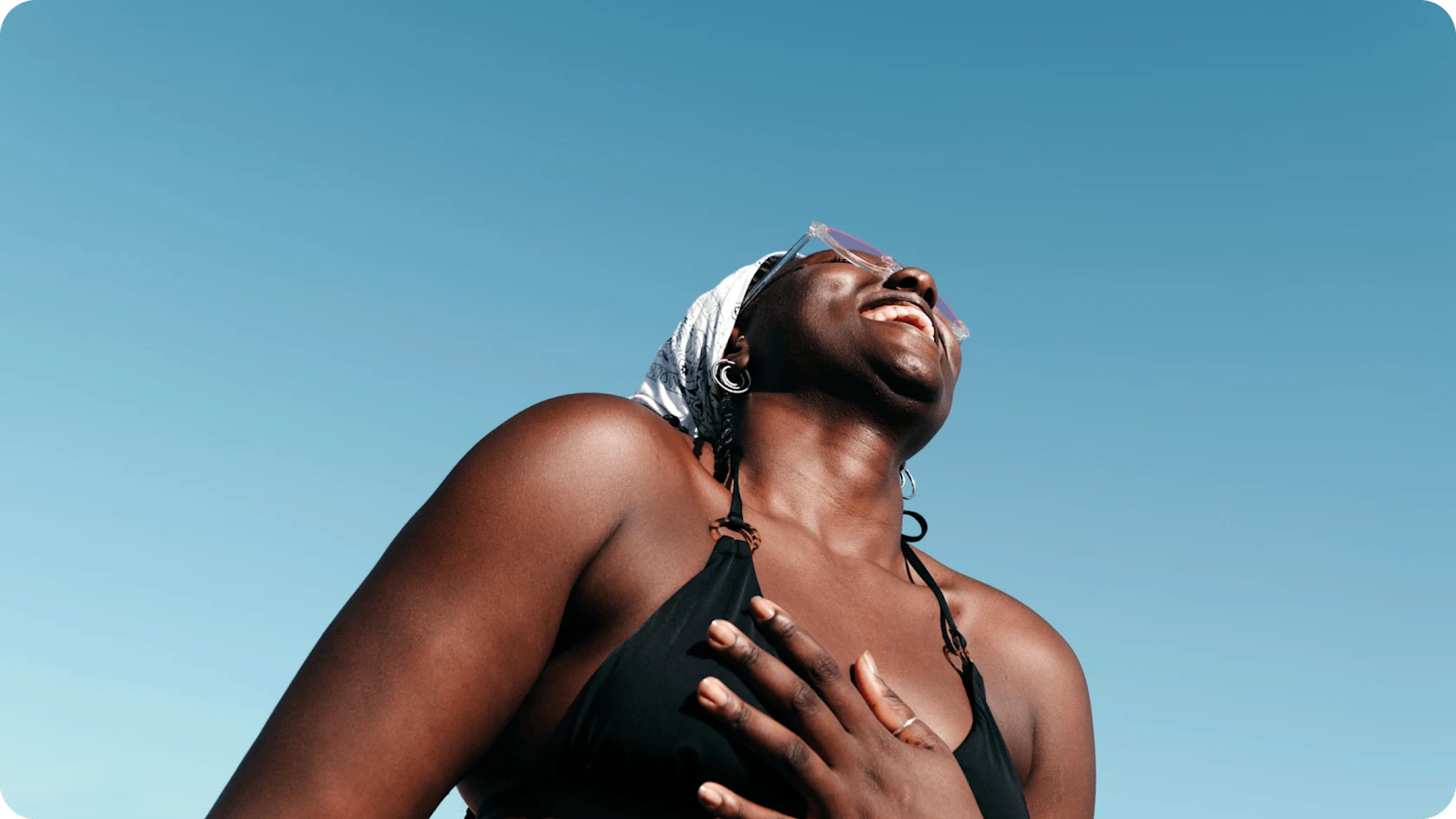 A woman in a bathing suit and sunglasses, smiling and looking up at a bright blue sky.