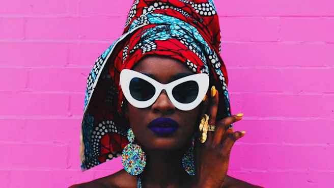 Un portrait d’une femme noire cadré au-dessus de ses épaules, avec un mur de briques rose vif en fond. La femme porte du rouge à lèvres violet, des lunettes de soleil blanches, un foulard rouge et bleu, et de gros bijoux.