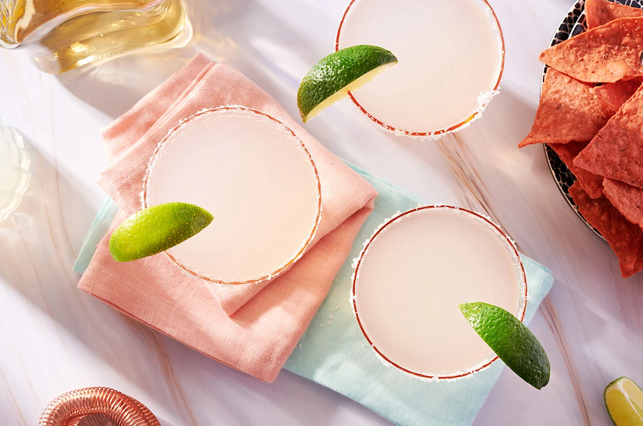 Overhead view of three margaritas garnished with lime wedges on pastel napkins, next to a plate of red tortilla chips, evoking a fresh summer party vibe.
