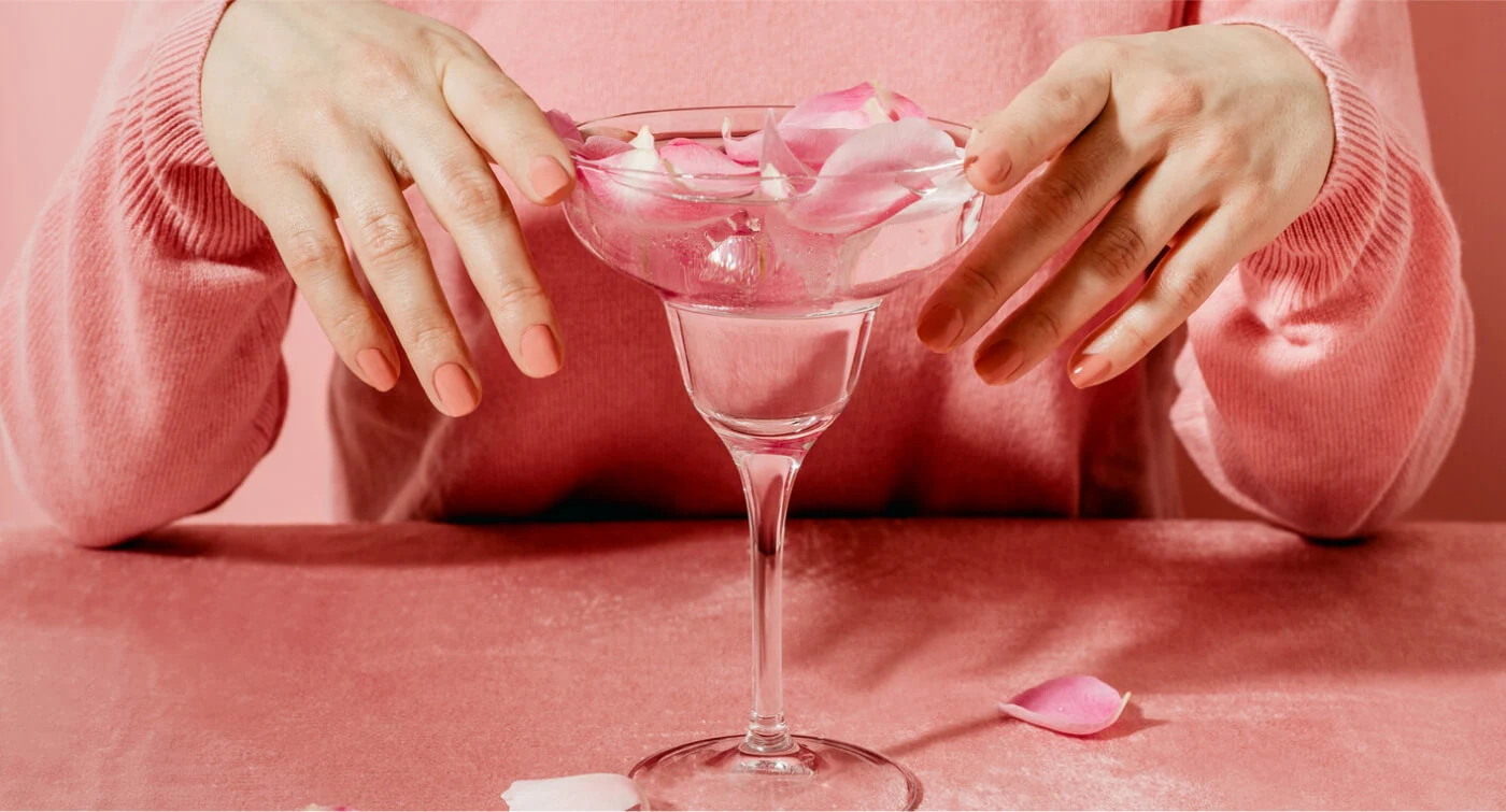 Une personne vêtue d’un pull rose clair et portant du vernis à ongles rose est assise à une table rose. Elle tient un verre contenant un liquide rose et des pétales de rose.