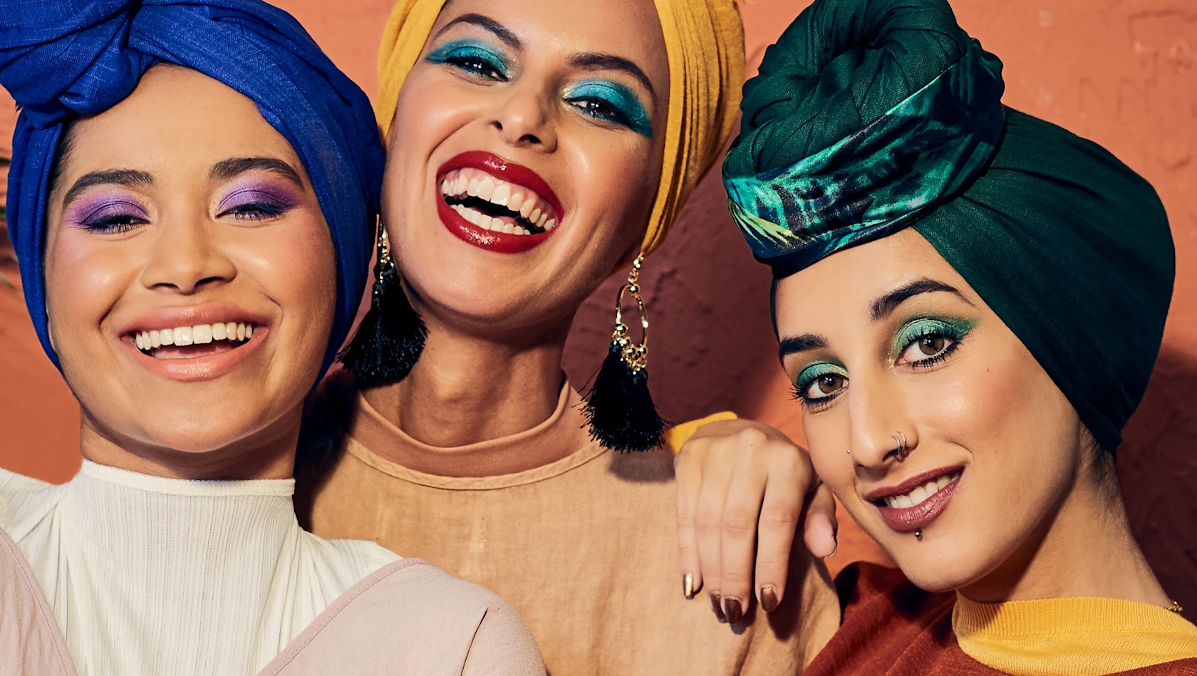Three smiling women, each dressed in a head scarf of a different color: deep blue, bright yellow and deep green.