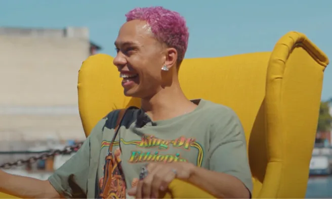 Young Gen Z man with pink hair sits in a yellow chair and laughs