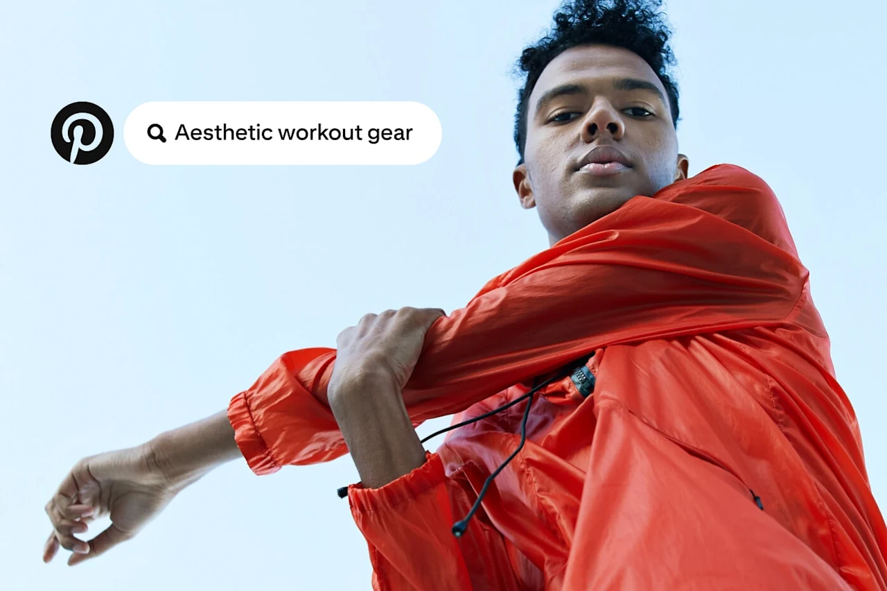 A person in a bright orange jacket stretches an arm while looking down at the camera. A Pinterest search bubble reads "Aesthetic workout gear."