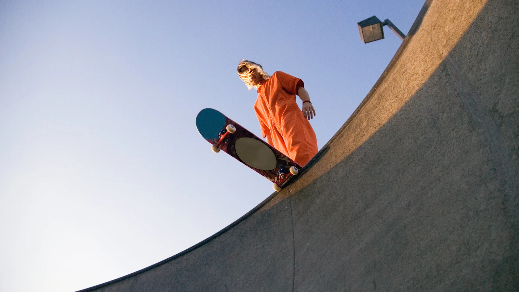 A White man with long blonde hair preparing to descend down a skateboard ramp on a pink and blue skateboard.