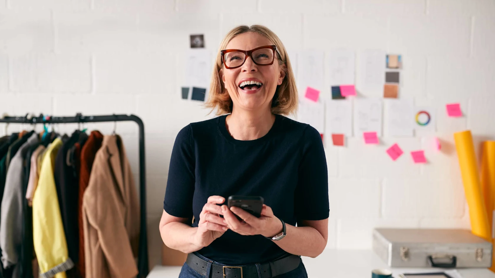 Older white woman with blonde hair and glasses looking up from her smartphone and laughing