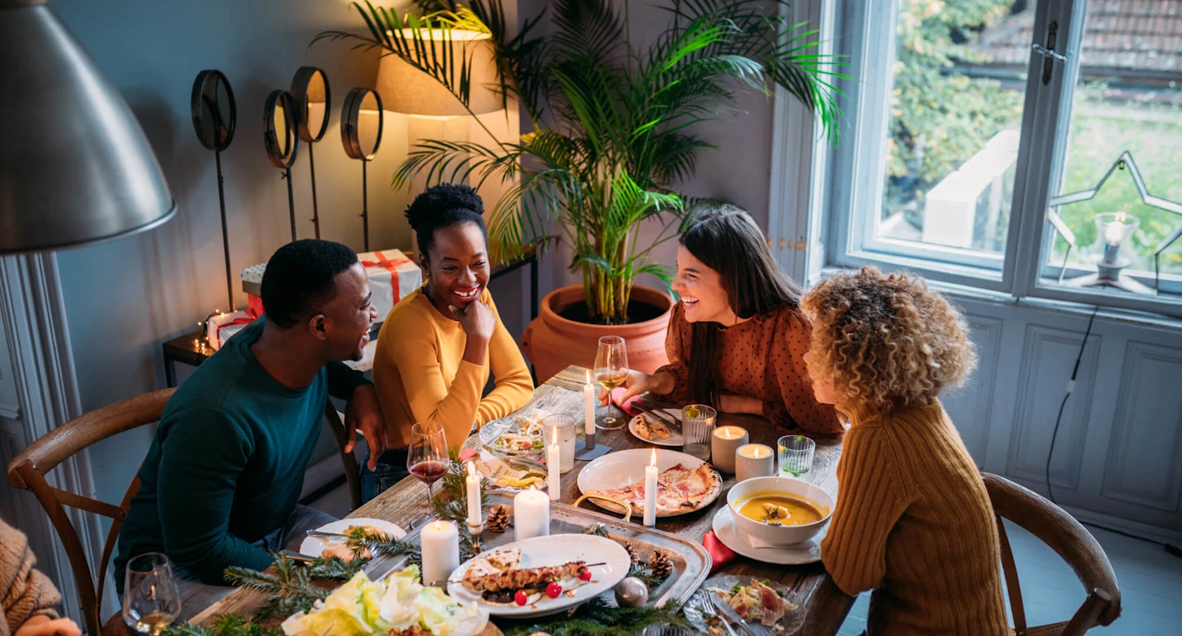 Four people are shown conversing around a dining room table. The table is set with various foods and decorative candles. It’s daytime outside, as shown out the window.
