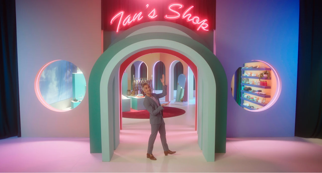 Tan France standing at the light green entrance of ‘Tan's Shop’. The shop's title is written in neon pink. Two circular windows appear to the left and right, giving viewers a glimpse of the shows and clothing items inside.