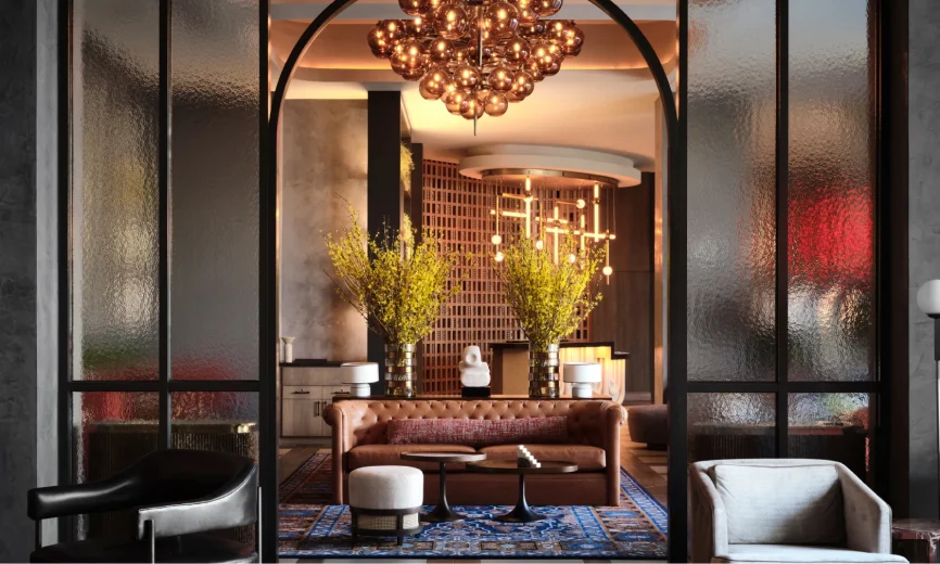 A Thompson Hotel lobby, featuring an arched entryway and a bronze chandelier. At the center of the room there's a leather couch, small ottoman and two coffee tables.