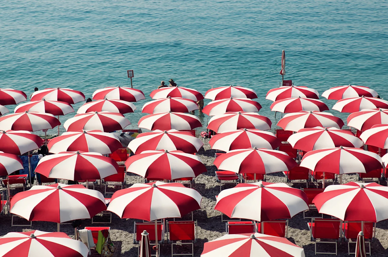 Rows of red and white beach umbrellas and chairs line a pebbled shore, overlooking bright blue water, capturing the energy of a sunny summer day by the sea.