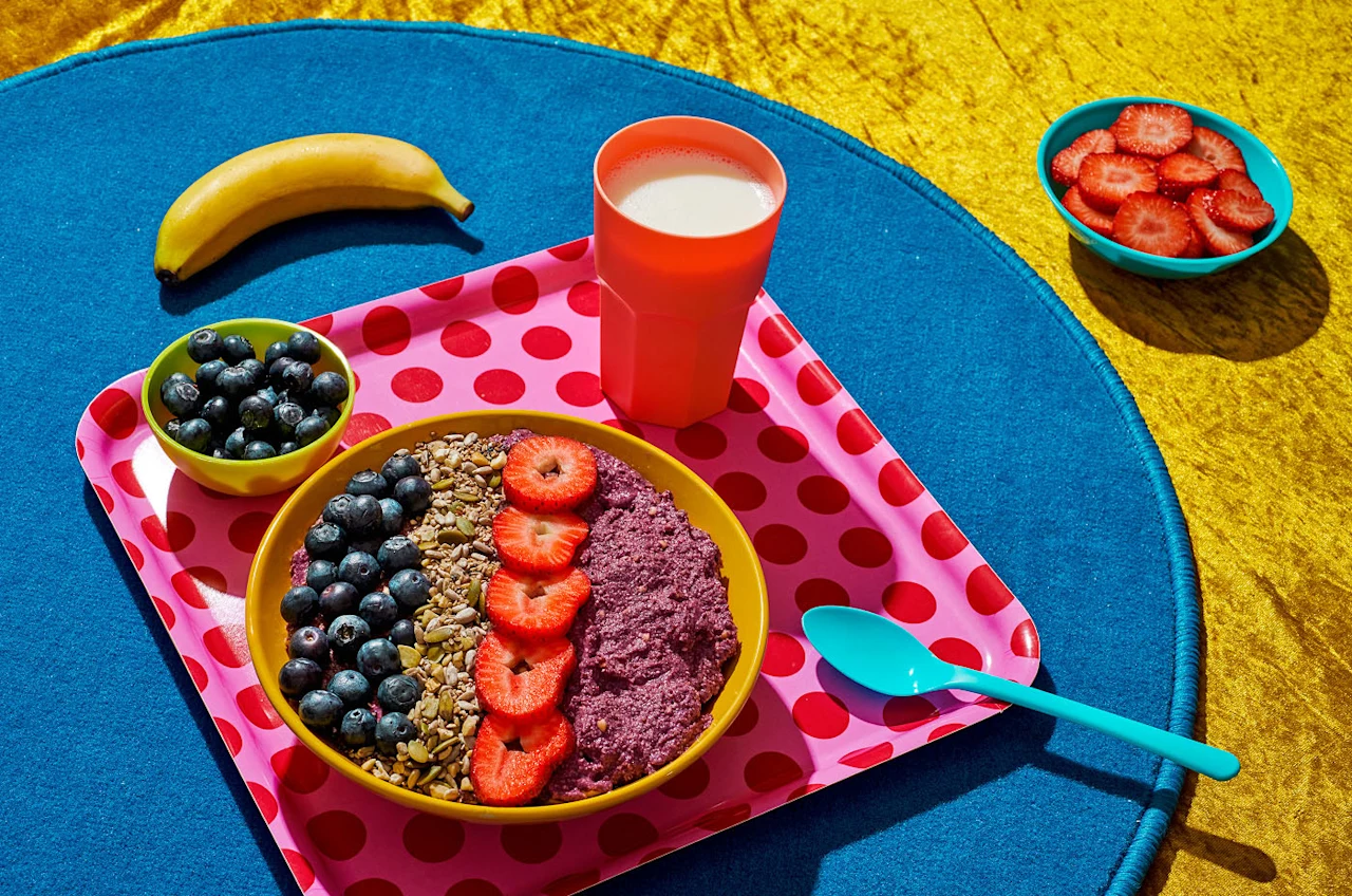 Colourful tray with a smoothie bowl topped with blueberries, granola and strawberries, a cup of milk, banana and bowls of fruit.