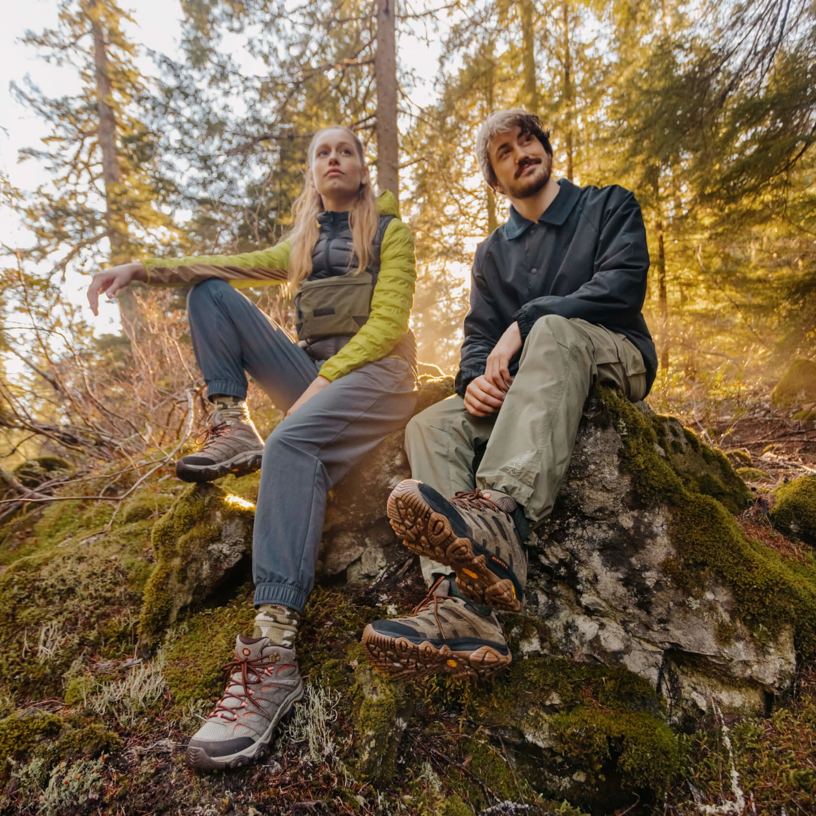 Young man and woman sitting outdoors on mossy rock, wearing outdoor gear and hiking boots