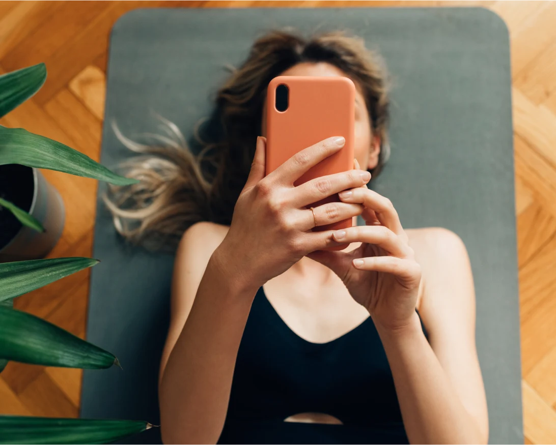 Просматривающая что-то на мобильном телефоне белая женщина, лежащая на сером коврике для йоги.