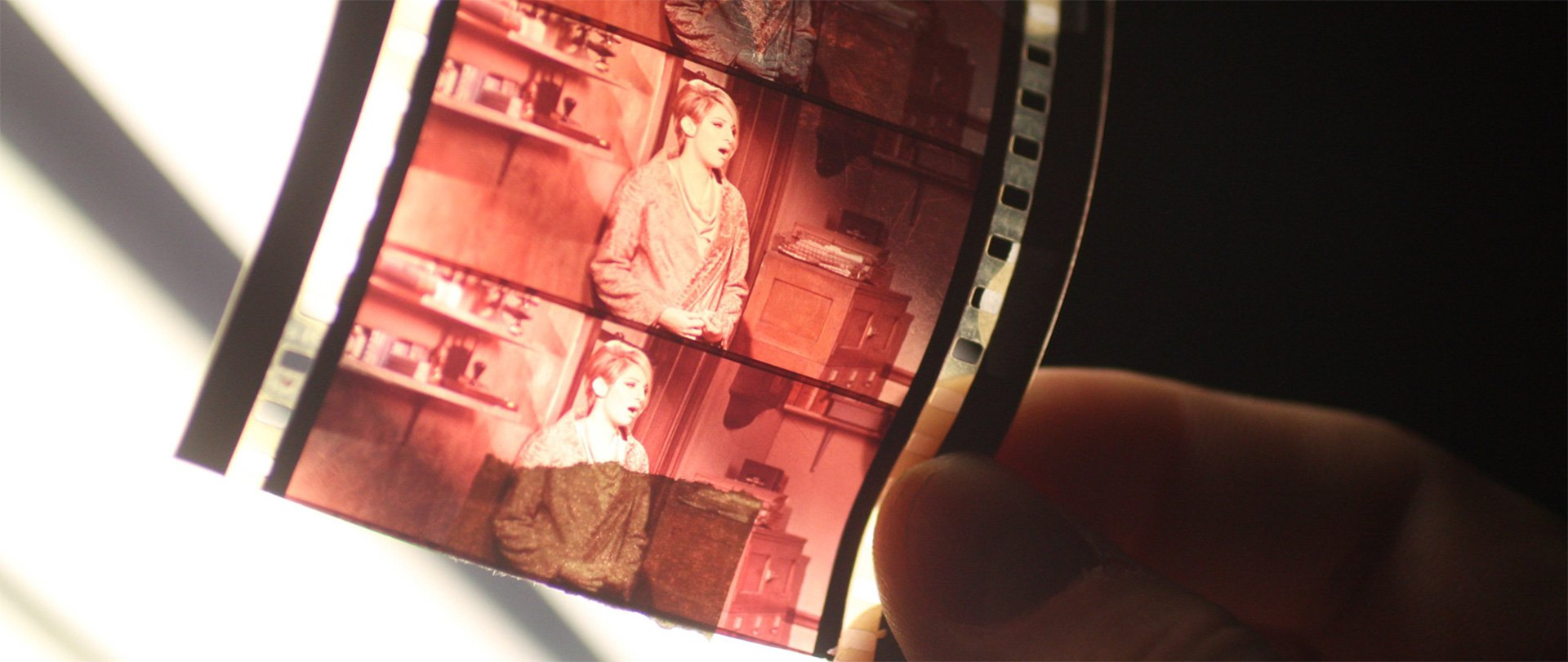 Närbild av en hand som håller upp en remsa rödtonad negativ film mot ljuset. På filmrutorna syns en person stående i ett rum med hyllor och möbler i bakgrunden. Bilden har mjukt fokus och varm belysning.