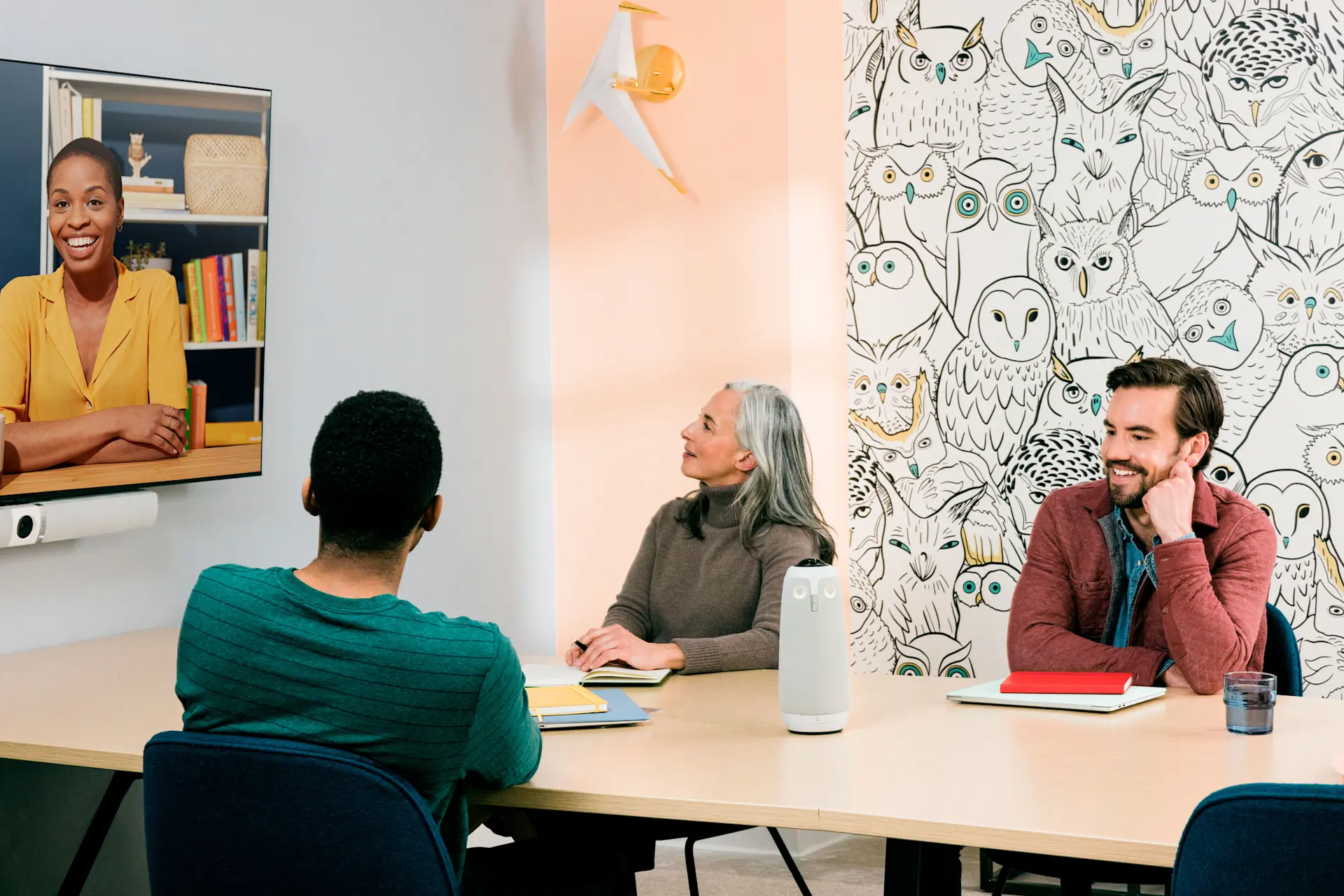A small group of co-workers engaging in a hybrid meeting and smiling, using the Meeting Owl and Owl Bar