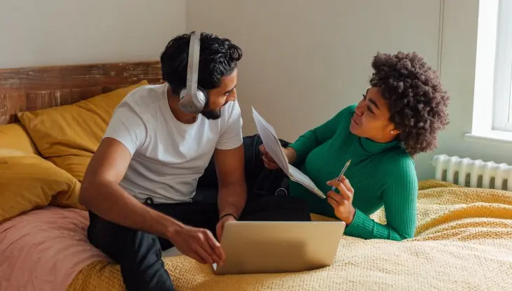 Couple with separate laptops looking at each other while sitting on a bed