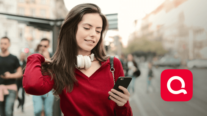 A woman wearing a red top along with white headphones is smiling while watching her mobile on the street.