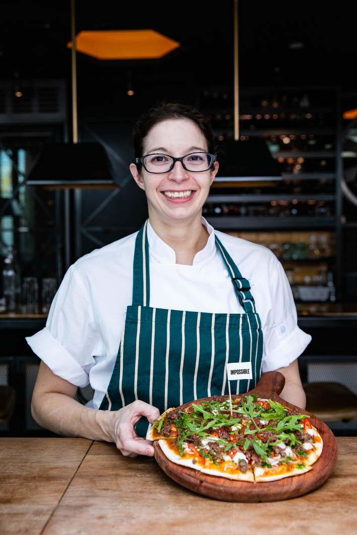 A woman chef smiling while serving Impossible™ Pizza with an Impossible™ flag and veggies in a restaurant.