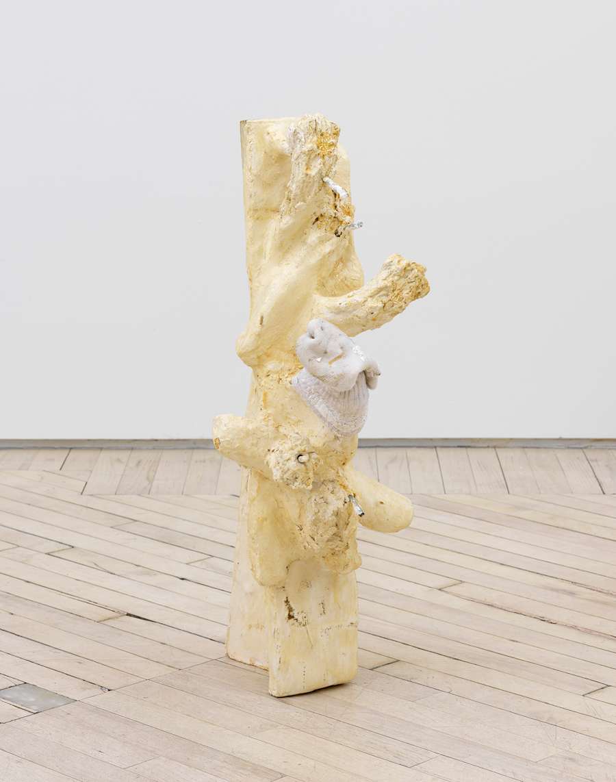 In a gallery space, an abstract irregularly shaped sculpture freestanding on the ground. The sculpture has a light-tan finish and multiple protrusions. One of the protrusions has a sock covering it.