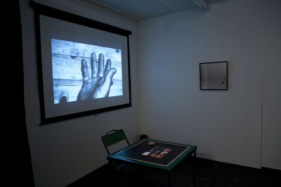 In a gallery space, a projection on the left wall displays a hand. In the middle of a room is a foldable chair and desk. A framed photograph is on a wall to the right of the table.