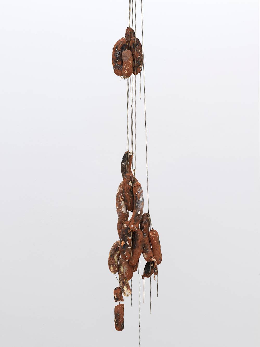 A sculpture suspended in a gallery space consisting primarily of sausage links hanging from chains.