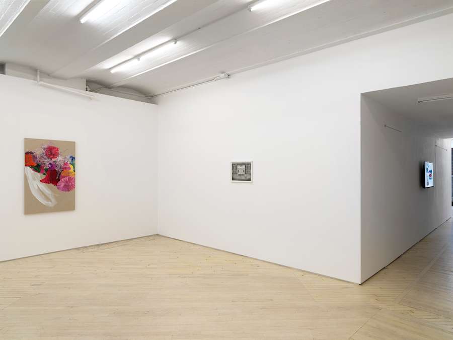 In a gallery space to the left a vertical painting on raw canvas depicting a group of bell shaped flowers in bright colors such as reds and pinks. To the right a small black and white drawing in a white frame. There is a long hallway with a darkened projection receding into the background.
