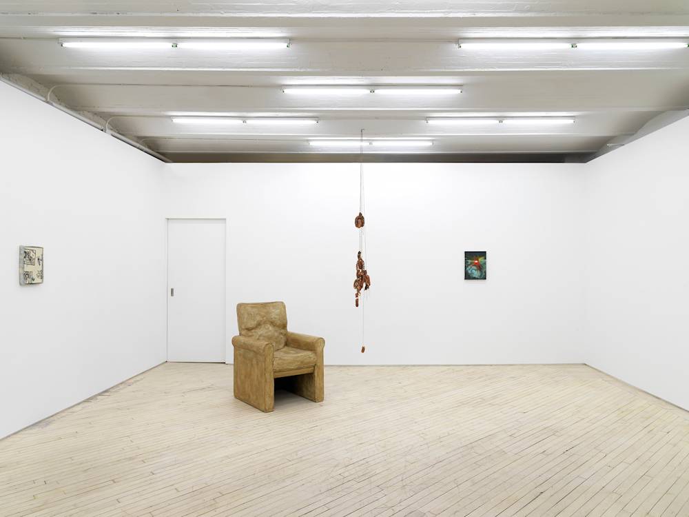 A gallery space with white walls and flourescent lights hanging from the ceiling. There is a doorway on the middle wall to the left. On the left wall is a small wall hanging piece that is white and blue. In the middle of the room is a large brownish sculpture depicting a chair. Next to the chair is a sculpture consisting of sausage links hanging from the ceiling. To the right is a small darkish-blue painting.