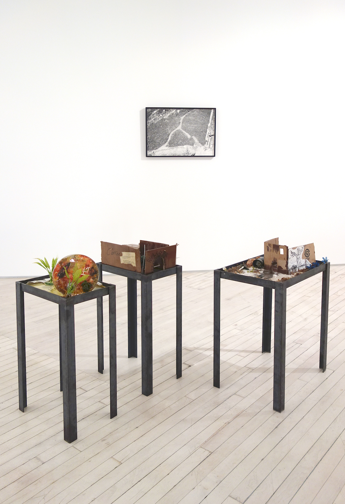 in the background, a framed b/w photograph of a view of a grassy and rocky field, in the foreground three table sculptures with hand made dioramas on top comprising fake green plants and cardboard boxes made into small, rough-hewn structures.