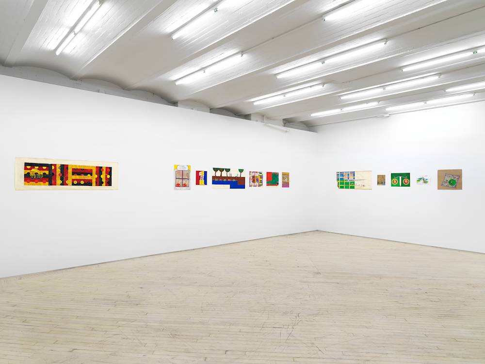 In a gallery space, a series of small works on paper are installed close together on a white walls. The drawings are clustered in groups. The drawings depict abstracted, cartoon-like character scenes. The sizes of the paper vary in length and proportion. There is a large scroll-like drawing rendered in the colors of the German flag.