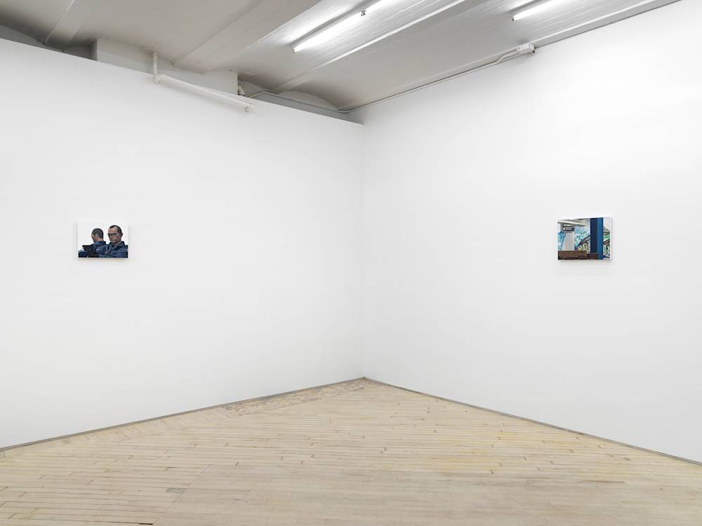 Angled view of the gallery with white walls, fluorescent lights and light wood floors. Two very small paintings hang on the 2 visible walls. At left is a portrait of a man with glasses who is holding a gun, and at right a colorful painting of a subway station with a mosaic of a fish.