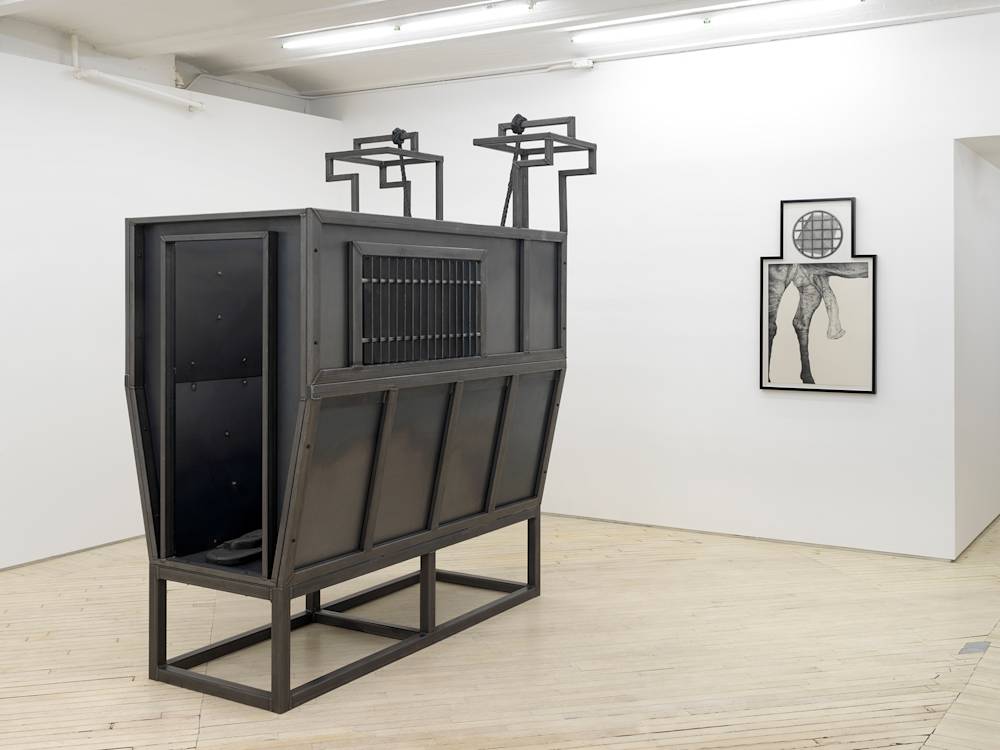 In a gallery space, a large floor iron floor sculpture resembling a cattle container. Behind the sculpture hung on the wall is a graphite drawing of horse genitals with a geometric pattern in a circle drawn above it in an irregularly shaped black frame.