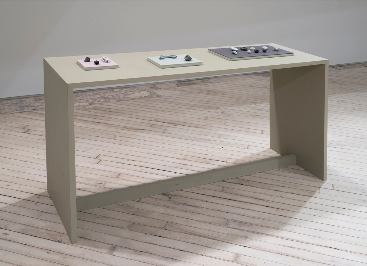 In a darkened gallery space, a green-tinted table. Resting on top of the table are three small plinths housing various miniature sculptural objects. The objects resemble bone forms or other organic matter.