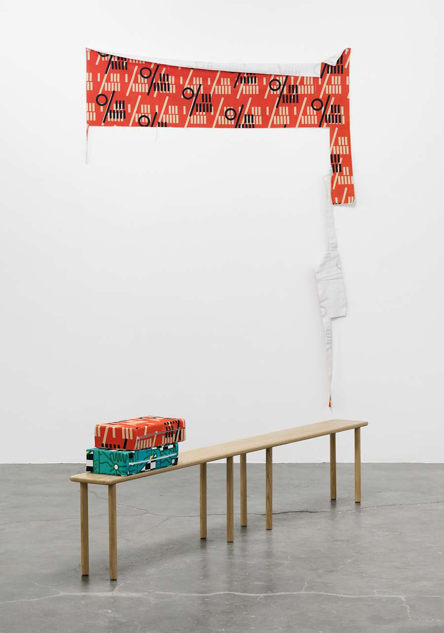 Installation view of Jaya Howey exhibition at Armada, Milan with a horizontal custom designed fabric swatch pinned to the wall allowing for some draping and a long wooden bench with eight legs and two custom fabric rectangular pillows on top.