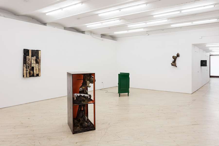 Installation view of an exhibition by Brandon Ndife. In a gallery there are two sculptures sitting on the floor appearing like pieces of furniture or cabinetry. Hanging on the wall are three other sculptures each appearing like a sconce, a piece of floor, and a kitchen cabinet.