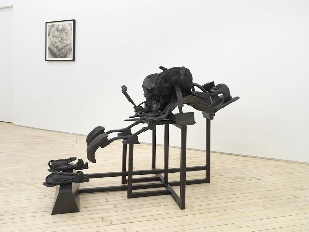In a gallery space, a large cast iron metal sculpture rests on the floor. The sculpture resembles a riding saddle and accompanying straps on top of an architectural bracing structure. To the left of the sculpture is a realistic graphite drawing of a detail of a human body part.