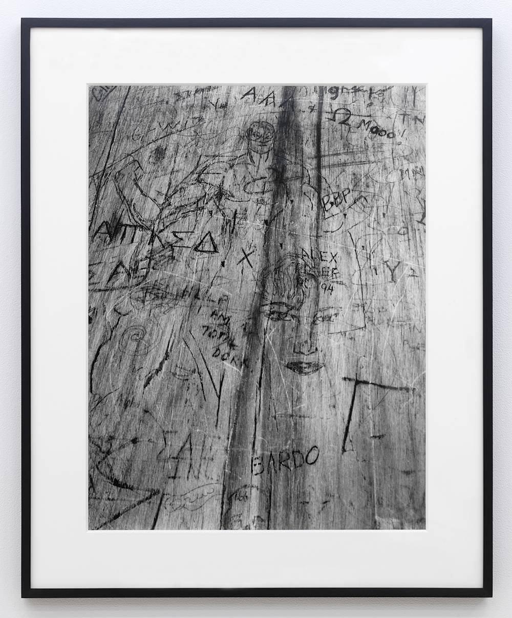 A black and white photograph depicting a bird's-eye view of a wooden desktop with numerous, carvings or marks throughout the surface. The photograph is in a black frame.