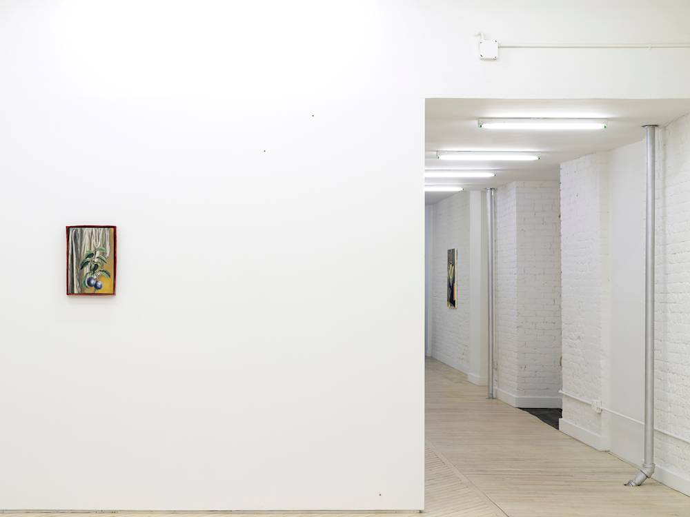 In a gallery space, a small painting depicting a bushel of blueberries in an irregularly shaped reddish frame. To the right of the wall is a long hallway with many fluorescent lights hanging from the ceiling. In the distance is another painting.