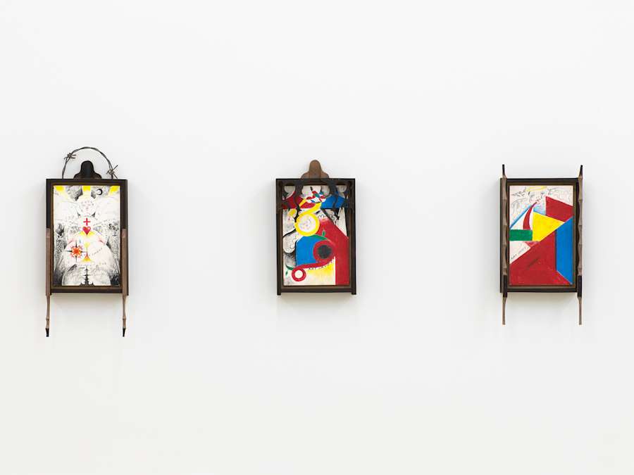 Installation view of three small vertical format drawings hanging on the wall all are framed in black wood with ornately carved frames. The first from the left also has barbed wire in an arc along the top edge of the frame. The drawing is black and white with accents of yellow and red, and shows concentric circles arranged geometrically, with rays of yellow sun at the top, symbols throughout, such as a red cross and red heart at center and a crescent moon and angel face which has the word 'Joy' written across the forehead. At left and right are two brick smokestacks. The two drawings on the right have solid irregular but geometric shapes in yellow, red, blue and green, as well as cartoonish angel heads with wings and haloes and the words 'Art Saves!' written in pencil.