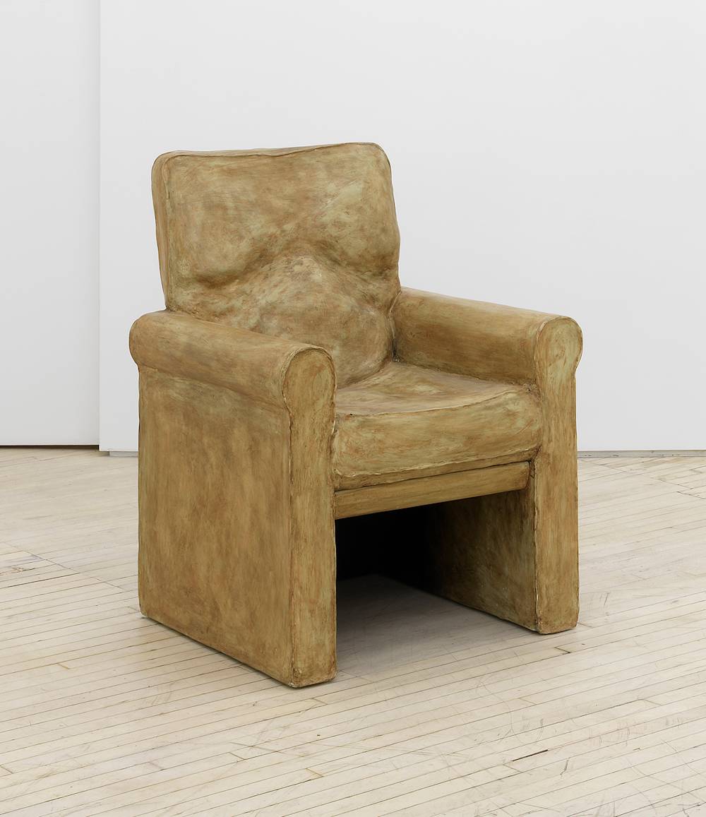 In a gallery space, a large brownish sculpture resembling a lounge chair. The finish is painted thin with brush marks throughout.