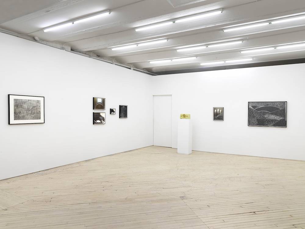 A gallery space with numerous framed objects throughout. On the left wall are a series of black and white framed images resembling photographs and drawings. On the right wall are two framed black and white photographs or drawings. In the corner is a doorway with a green vintage radio to the right of it on a pedestal.