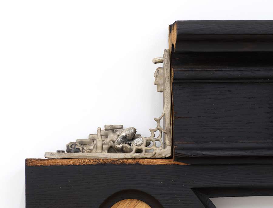 Detail view of top left of blackened wood archway sculpture. There is a square notch cut out of the wood, and in the niche is a metal L bracket that has a small tableau in cast bronze. A small webbing is at the angle joint and along the bottom is a pile of bricks, a reclining heart and a bottle, a profile of a head is on top right.