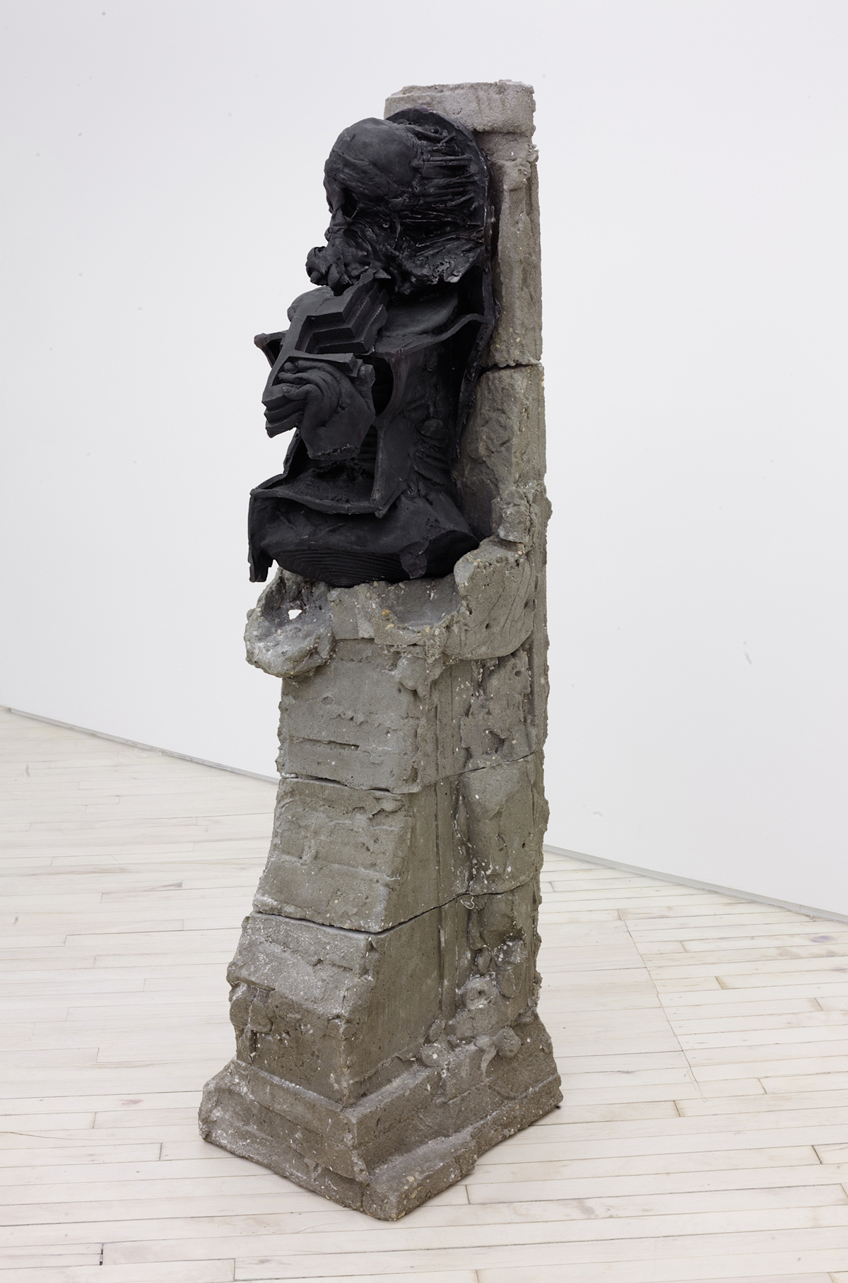 In a gallery space, a vertical metal and concrete abstract sculpture sits in front of a white wall.