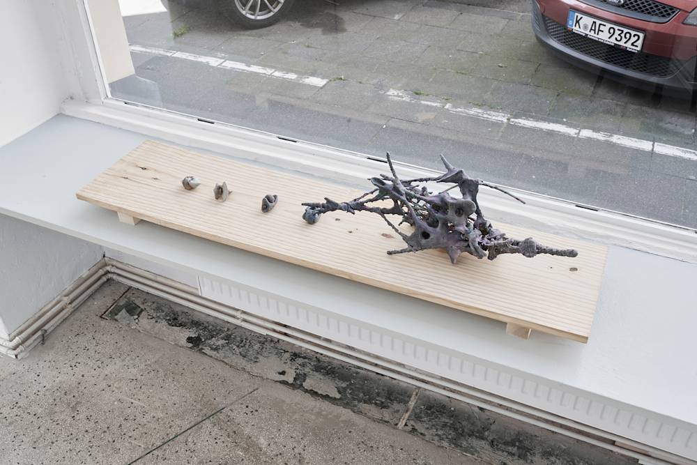 Four small hand-sized sculptures and one medium scale sculpture are presented on a rectangular piece of wood in front of a large window that opens up to a street view. The sculptures are amorphous, skeletal, and gray. They are resting on a bench inserted between two walls.