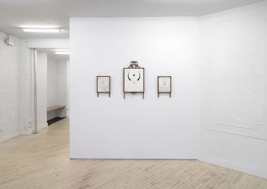 Image of three framed drawings, two small drawings flanking one larger drawing, all framed in wood and hanging on a white wall with a hallway on the left side of the image.