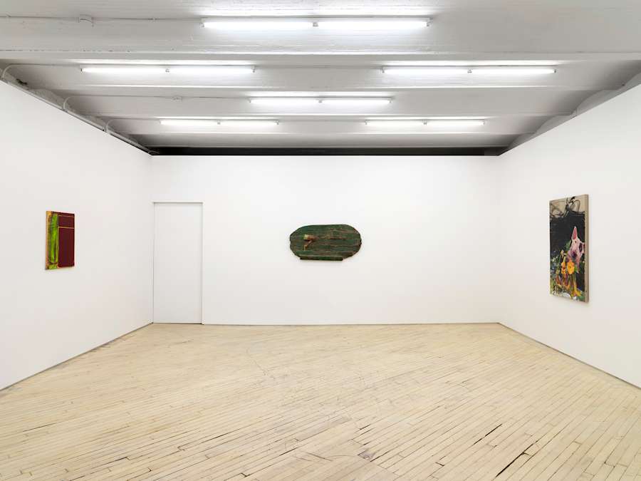 In a gallery space, a redish-green abstract painting is hung to the left. In the center a greenish wall hanging sculpture. To the right is an abstract painting with the head of a dog partially in view.