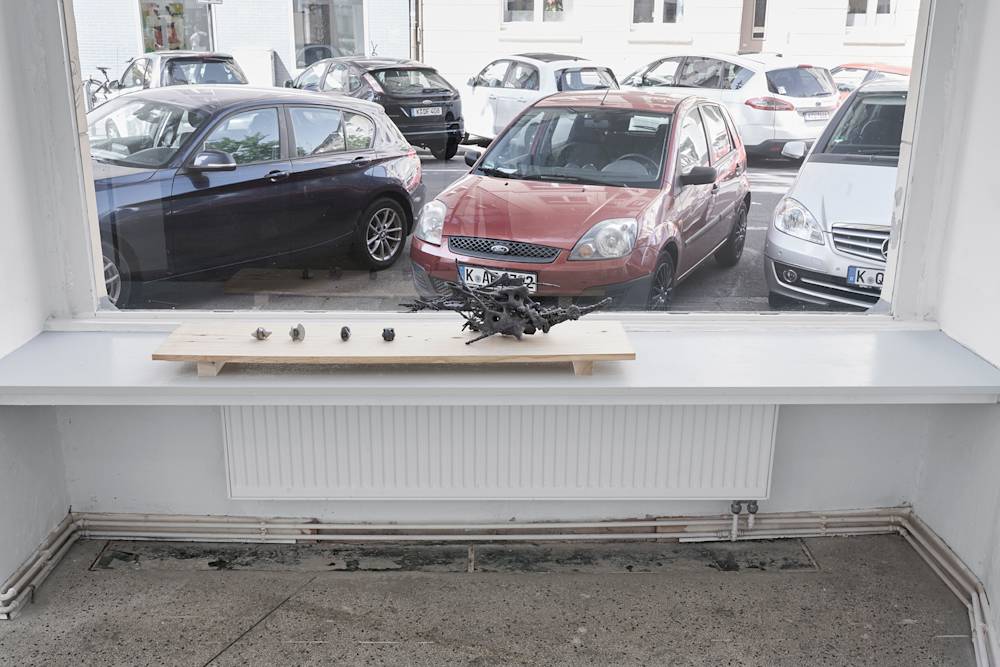 Four small hand-sized sculptures and one medium scale sculpture are presented on a rectangular piece of wood in front of a large window that opens up to a street view. The sculptures are amorphous, skeletal, and gray. They are resting on a bench inserted between two walls.