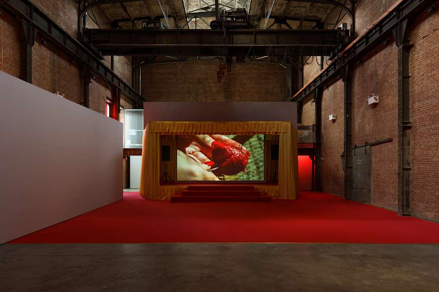 A darkened brick gallery space with an installation resembling a red carpeted concert stage with yellow curtains. In the center of the stage is a large video projection of a person cutting into a red piece of fruit.