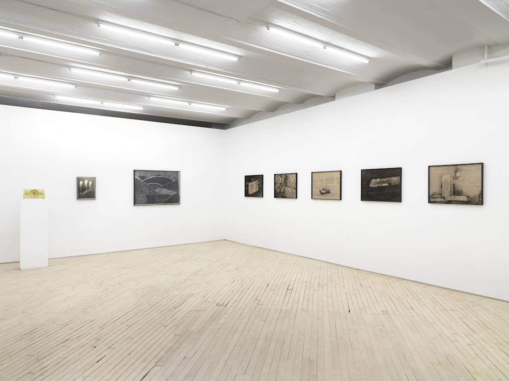 A gallery space with numerous framed objects throughout. On the left wall are two framed black and white photographs or drawings. In the corner is a green vintage radio on top of a pedestal. On the right wall is a series of pencil drawings behind tinted glass frames.