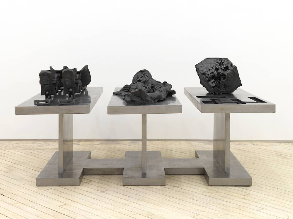In a gallery space, a large cast iron metal sculpture rests on the floor. The sculpture is made up of multiple components. Cast iron forms resemble a contraption device, an abstracted body, and a cubic form.